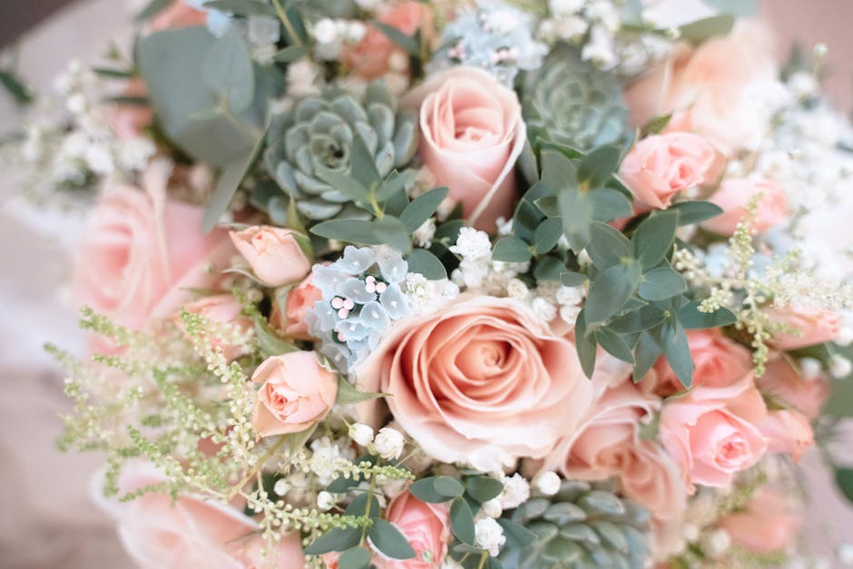 A close-up view of a floral bouquet featuring large, soft pink roses surrounded by smaller pink rosebuds, complemented by pale blue succulents and delicate white baby's breath. The arrangement is ador