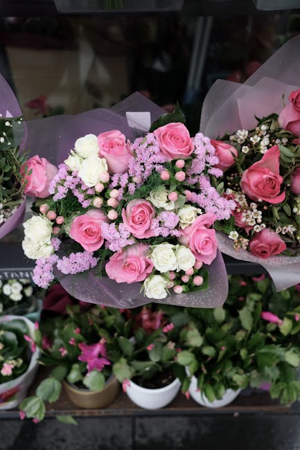 A beautifully arranged floral bouquet featuring pink roses, white carnations, and small pink and white filler flowers, wrapped in light purple tissue paper and tied with a ribbon, displayed on a store